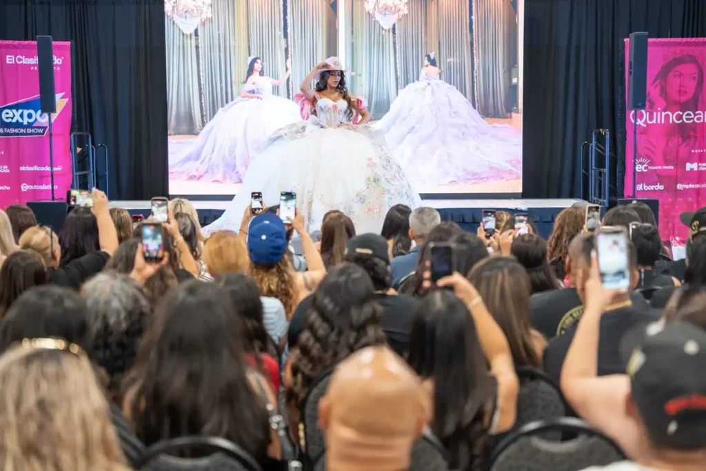 Quinceañera fashion show at the Quinceanera.com Expo in Ontario, California, as attendees photograph a model in a white gown.